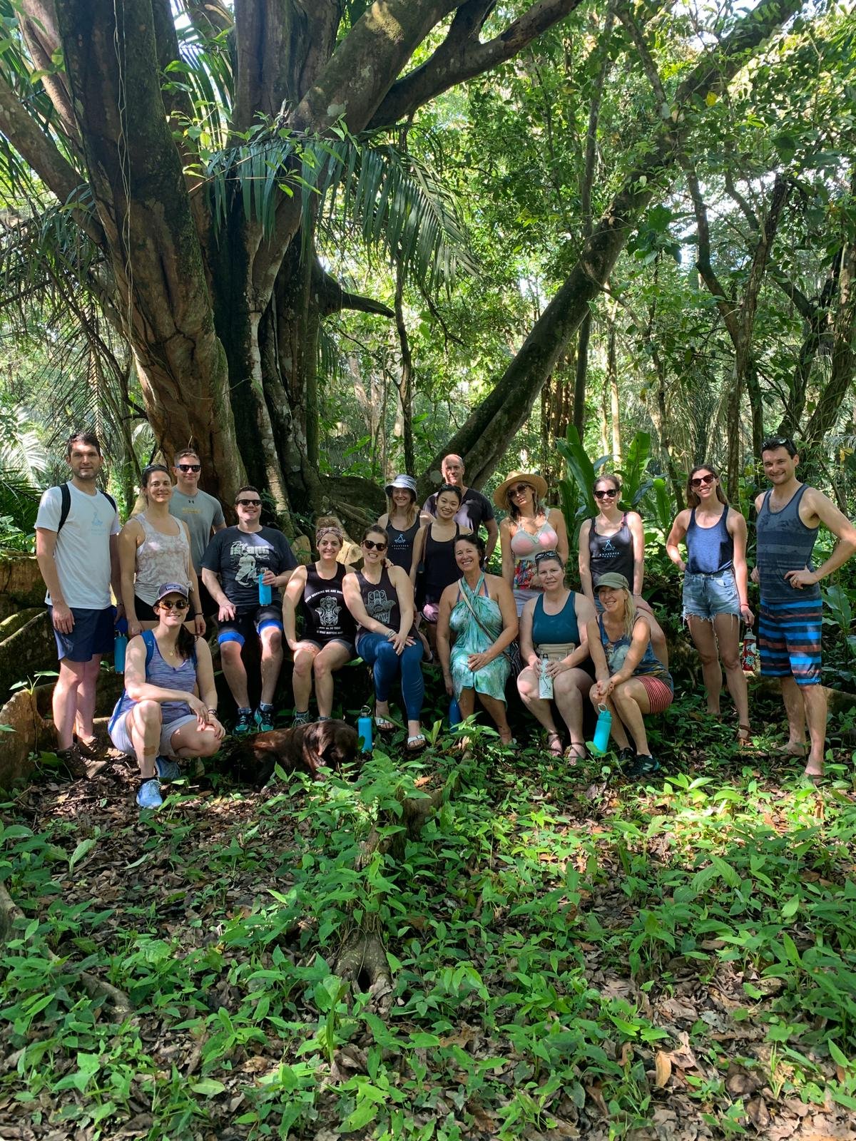 About eighteen retreat participants pose in front of a massive ancient tree on a jungle hike at Blue Osa, with a dog nearby and dense green foliage in the background.