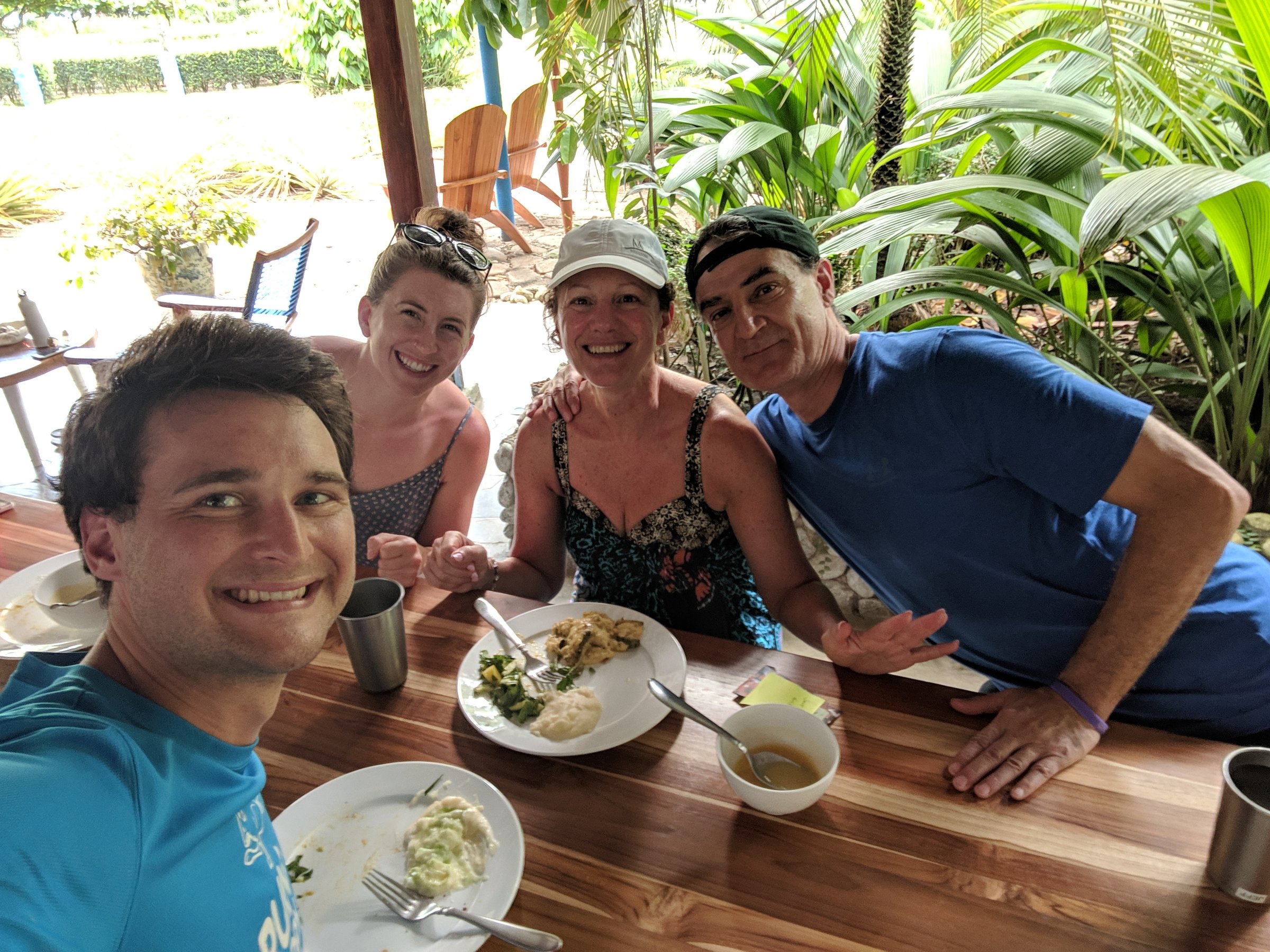 Four retreat participants smile for a selfie at a wooden dining table with plates of food, with bright tropical greenery behind them.