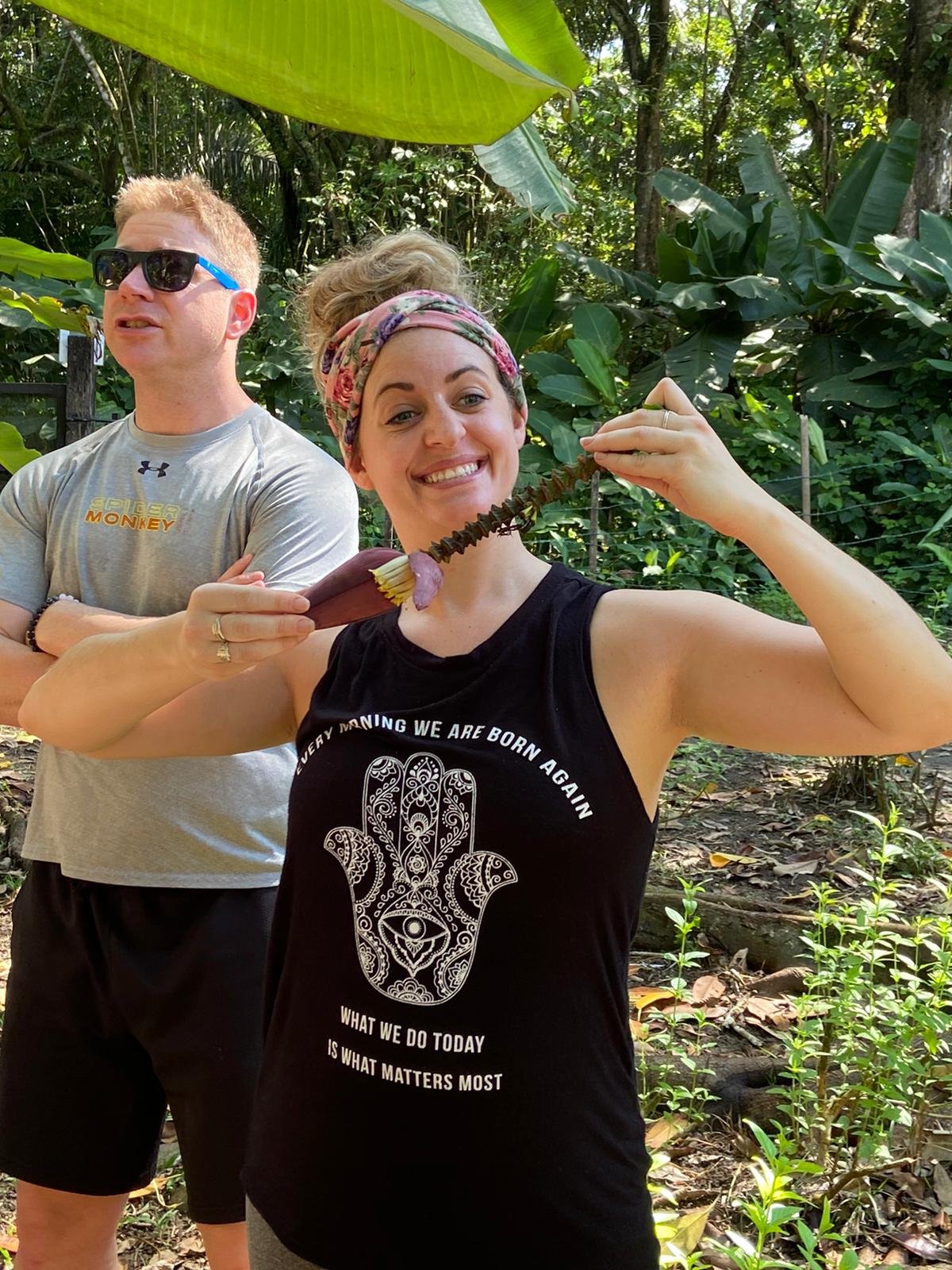 A smiling woman in a Hamsa-hand tank top and floral headband holds a banana blossom stalk in the jungle while a man in a Spider Monkey shirt stands behind her among broad tropical leaves.