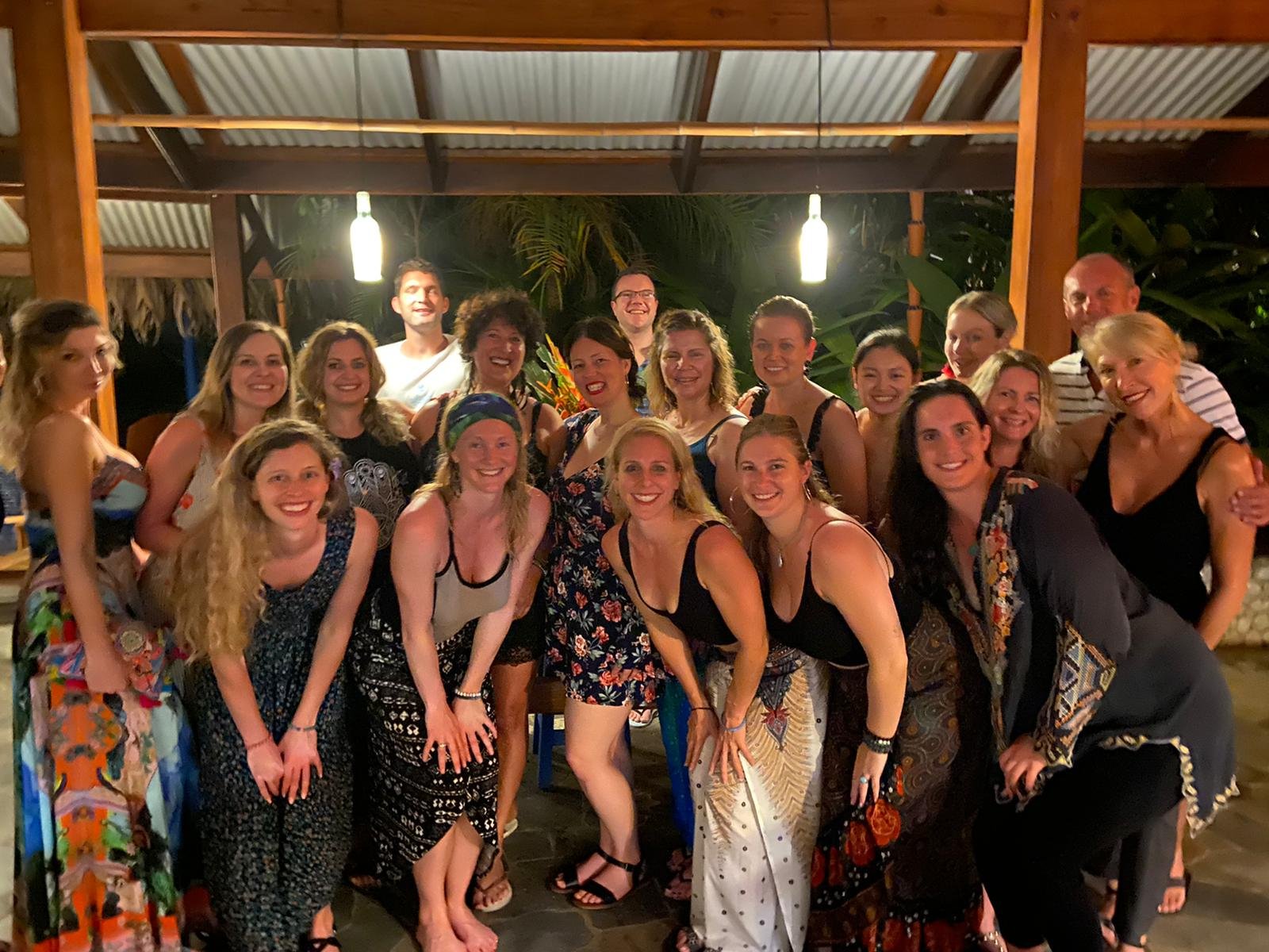 About eighteen retreat participants in colorful summer clothes pose together under a wooden pavilion at night at Blue Osa, smiling beneath bottle pendant lights with dark tropical foliage behind them.