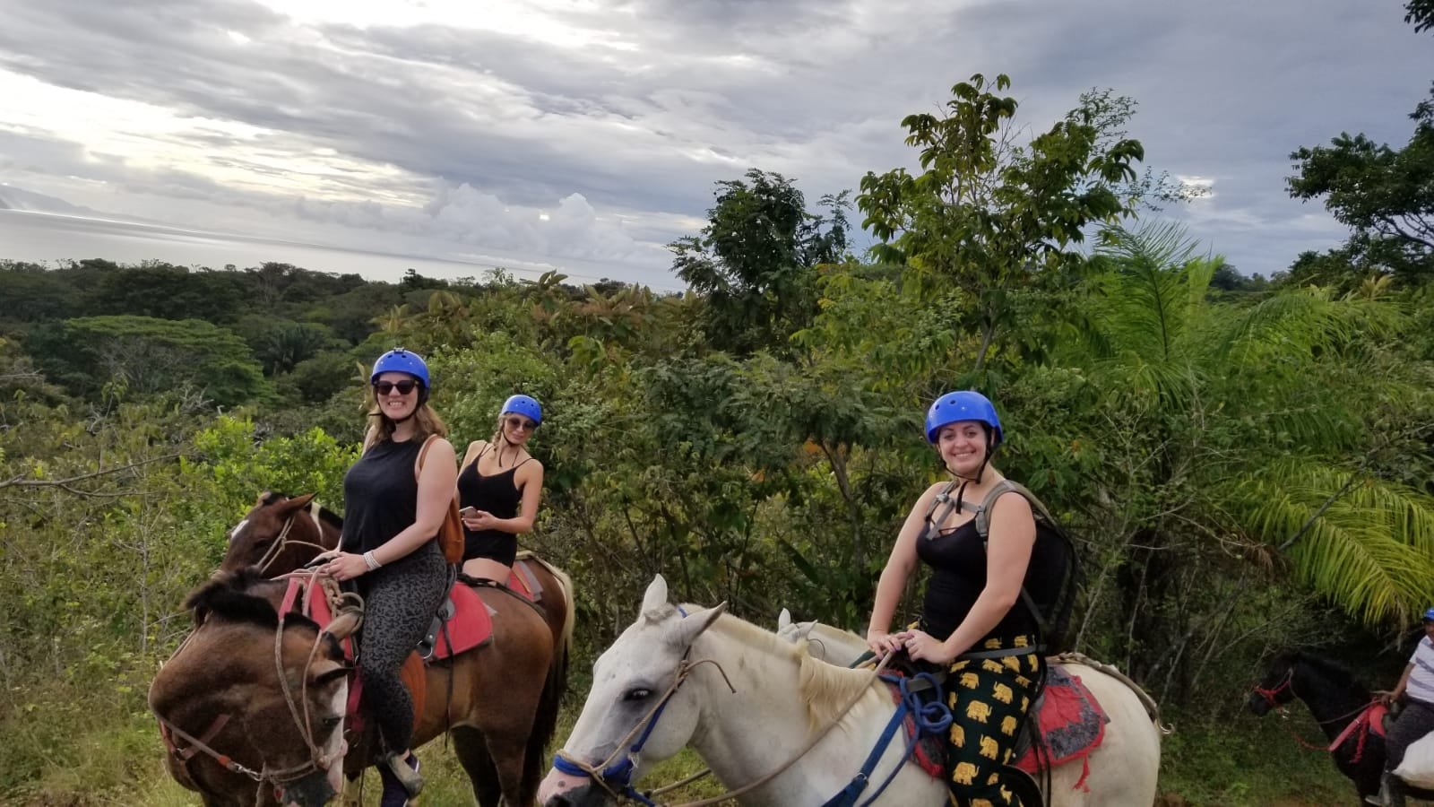 Three women in blue helmets and black tank tops smile while horseback riding brown and white horses on a lush jungle trail with the ocean visible in the distance under cloudy skies.