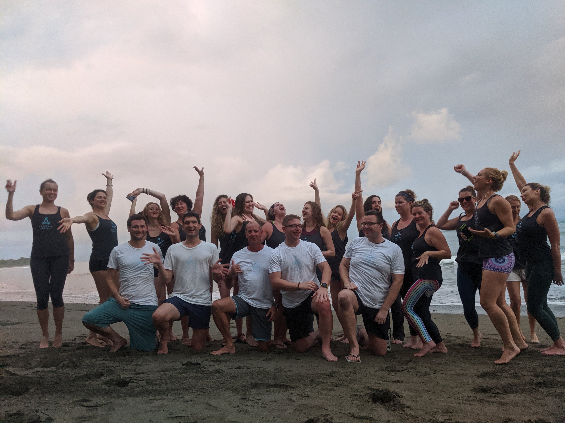 About twenty joyful retreat participants in matching black tank tops and white shirts pose on dark sand at the beach, many with arms raised, with gentle waves and an overcast sky behind them.
