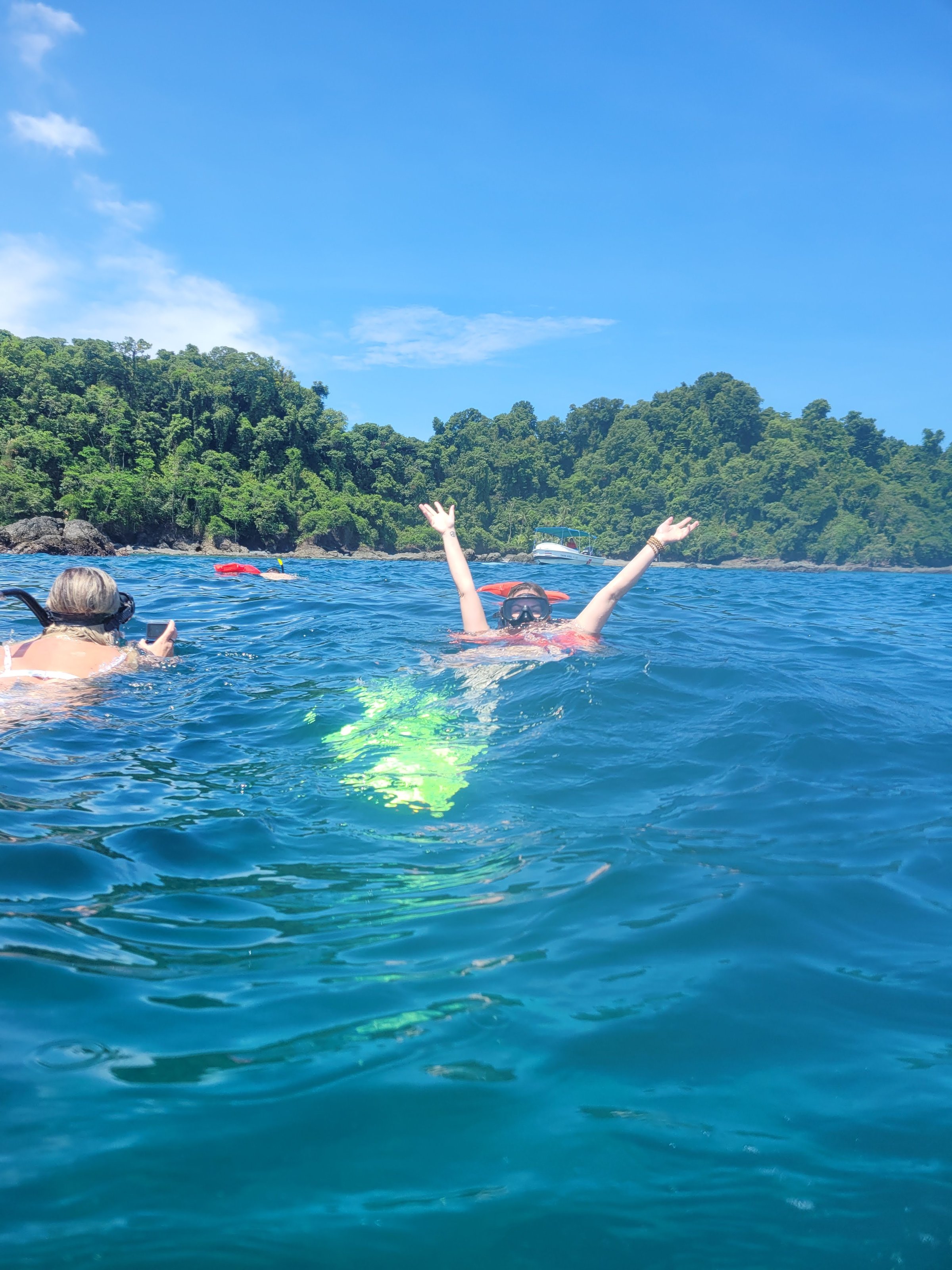 A snorkeler in a mask and pink life vest raises both arms in celebration in deep blue water with forested hills and sky behind.