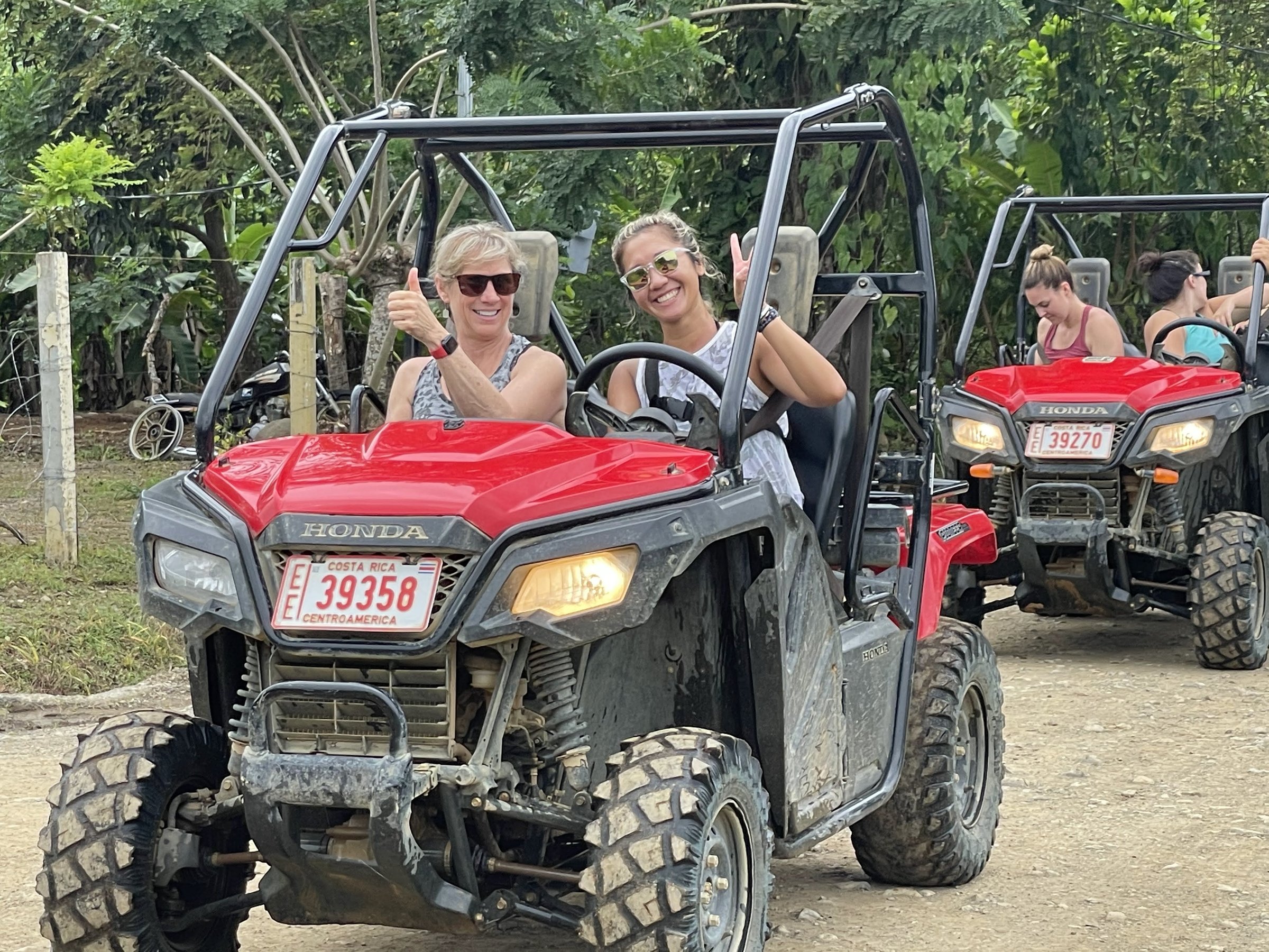 Two smiling women in sunglasses give a thumbs-up and peace sign from the front seats of a red Honda UTV on a dirt road through tropical forest.