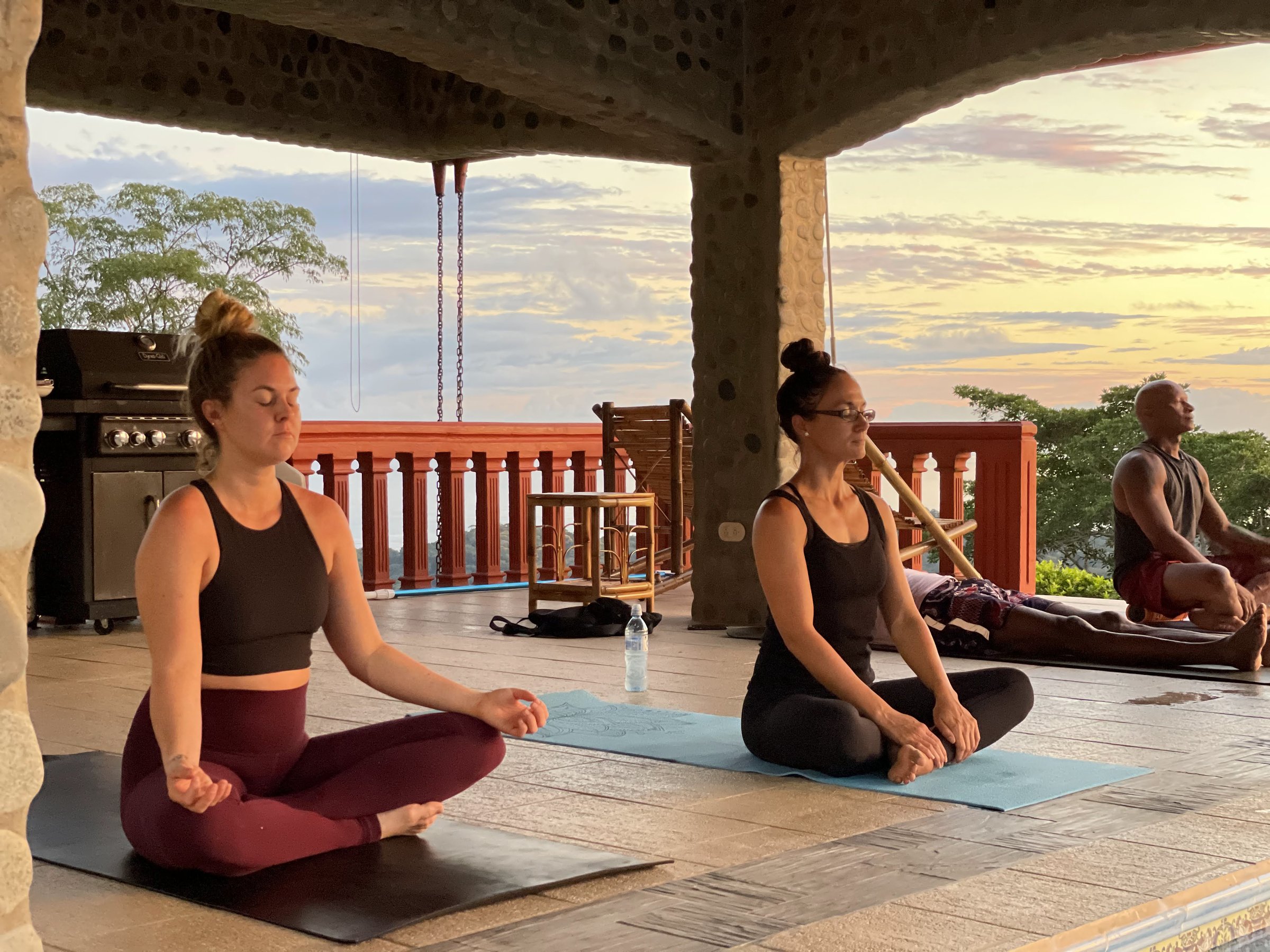 Three women meditate in cross-legged poses on yoga mats on an open terrace at sunset with orange sky and tropical trees beyond a red railing.