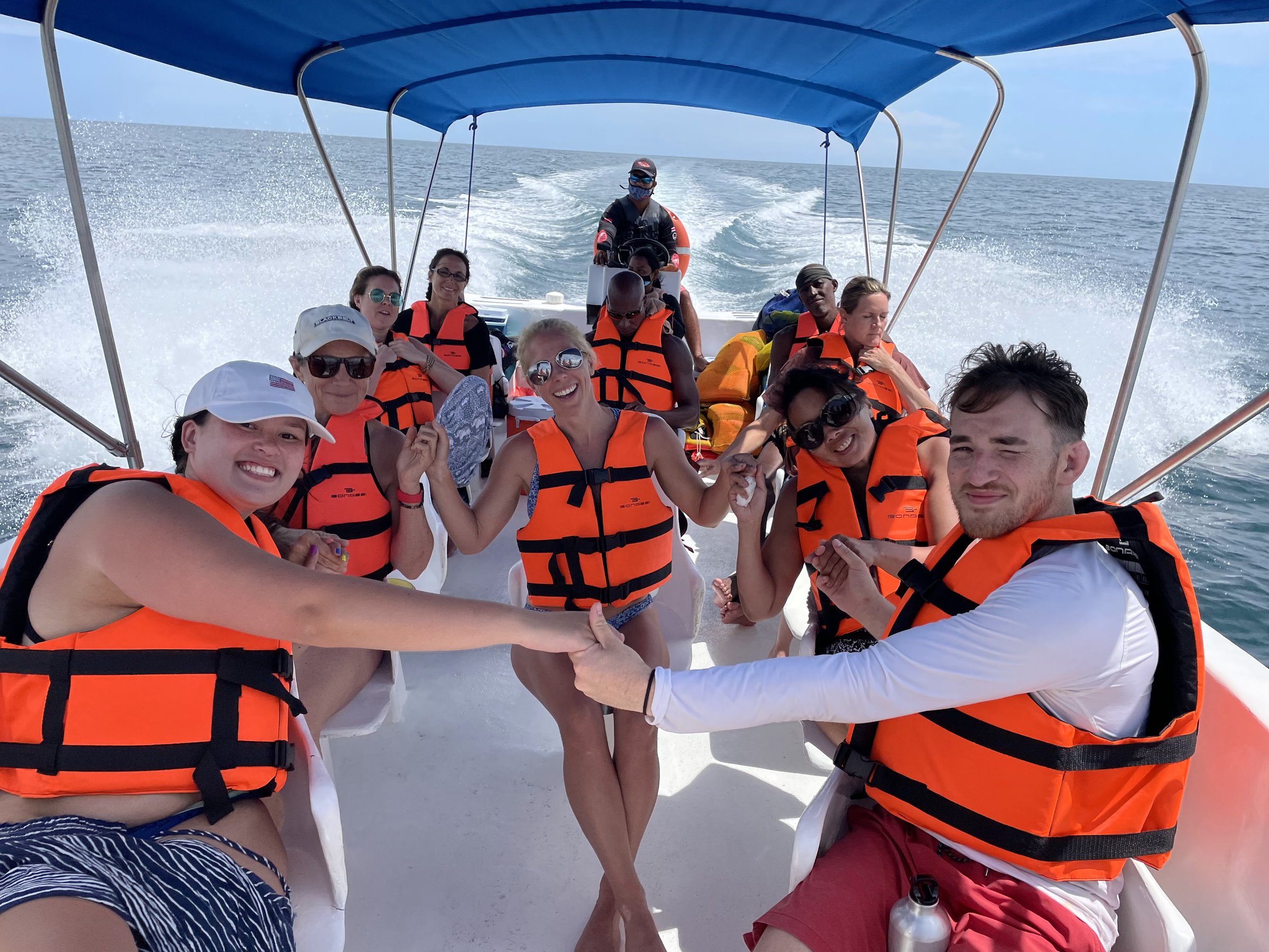 Retreat participants in orange life vests smile on a motorboat crossing turquoise water under a blue canopy with a white wake behind.