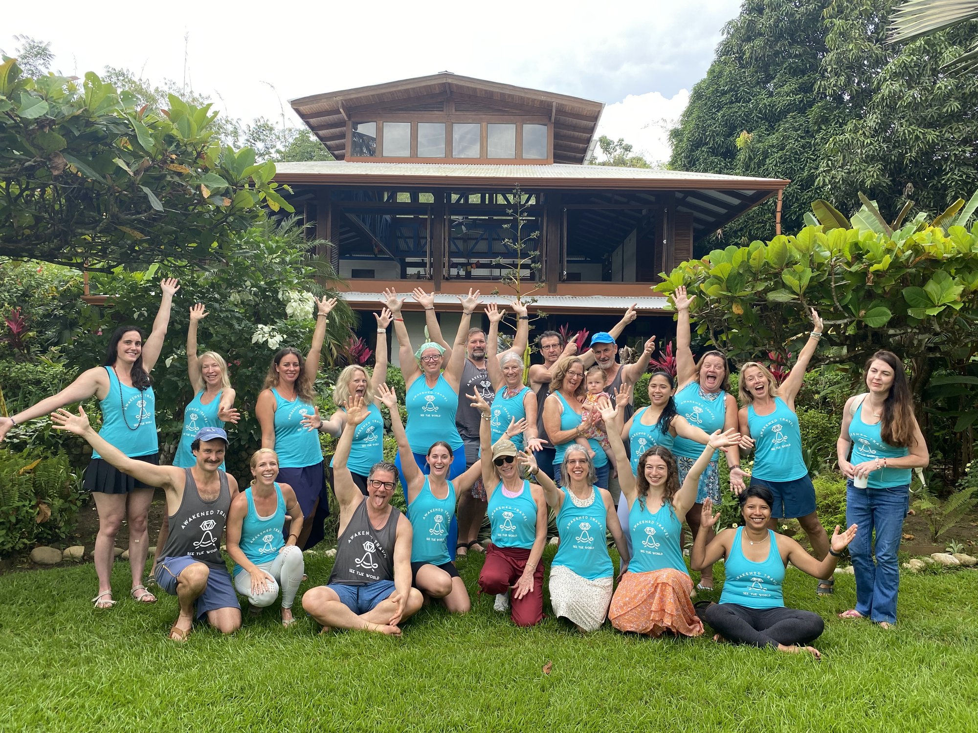 About twenty retreat travelers in matching teal Awakened Travel shirts pose on a green lawn in front of the wooden Blue Osa lodge, many with arms raised.