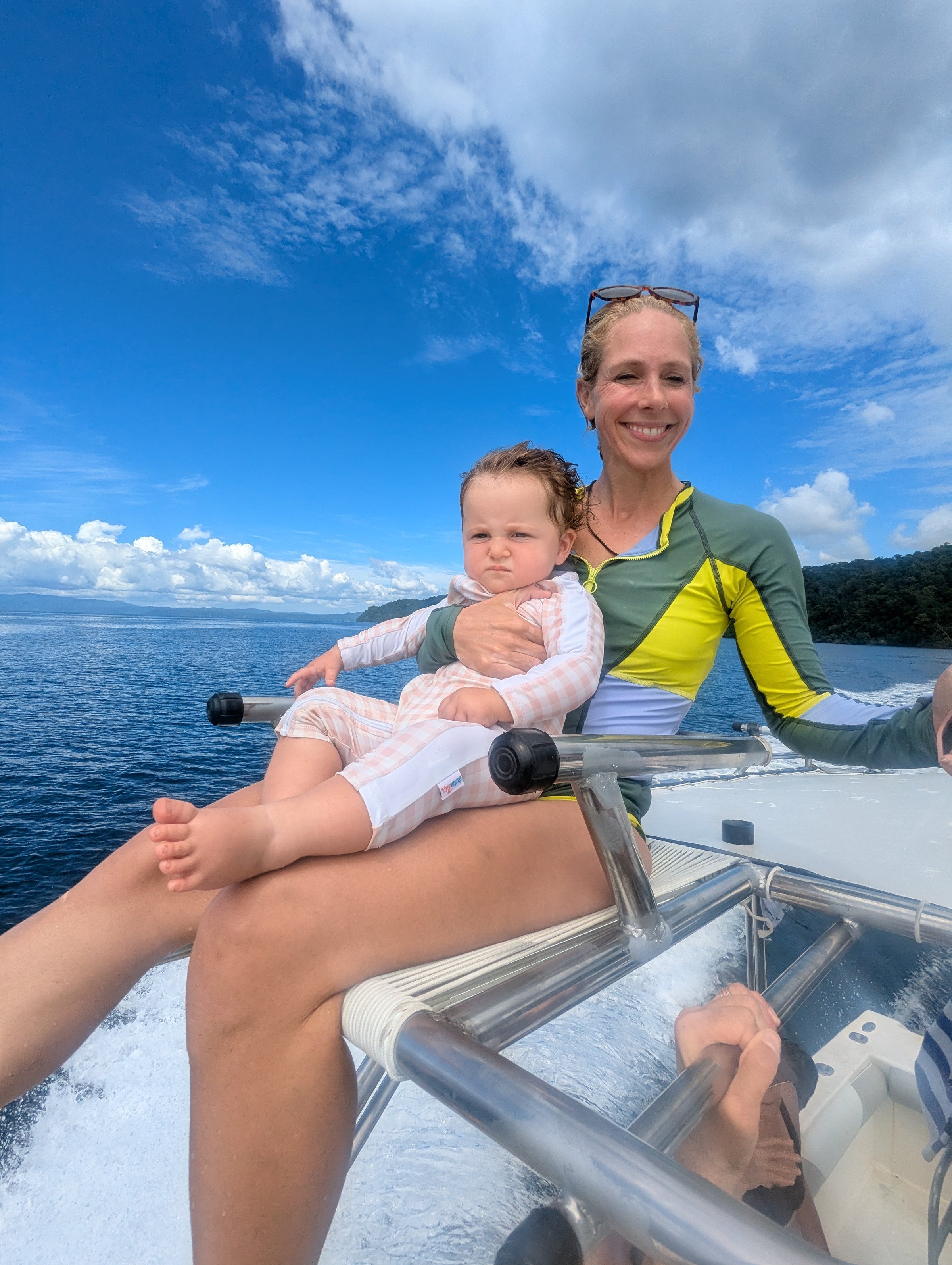 A smiling woman holds a baby on a boat railing as blue ocean and a green coastline stretch behind them on a sunny day.