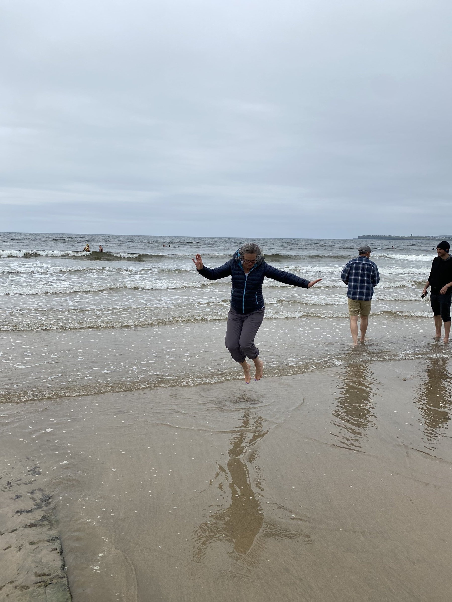 A woman in a blue puffer jacket jumps joyfully on a sandy beach with gentle waves and other people nearby.