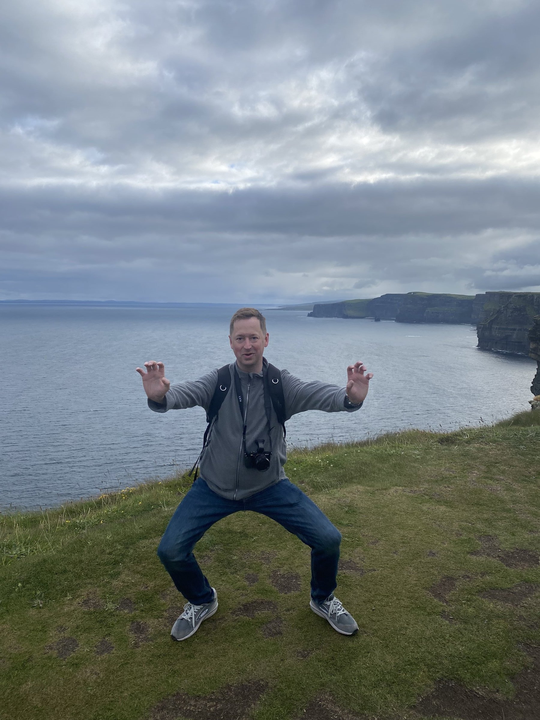 A man strikes a playful crouched pose on a grassy clifftop overlooking the ocean and distant cliffs.