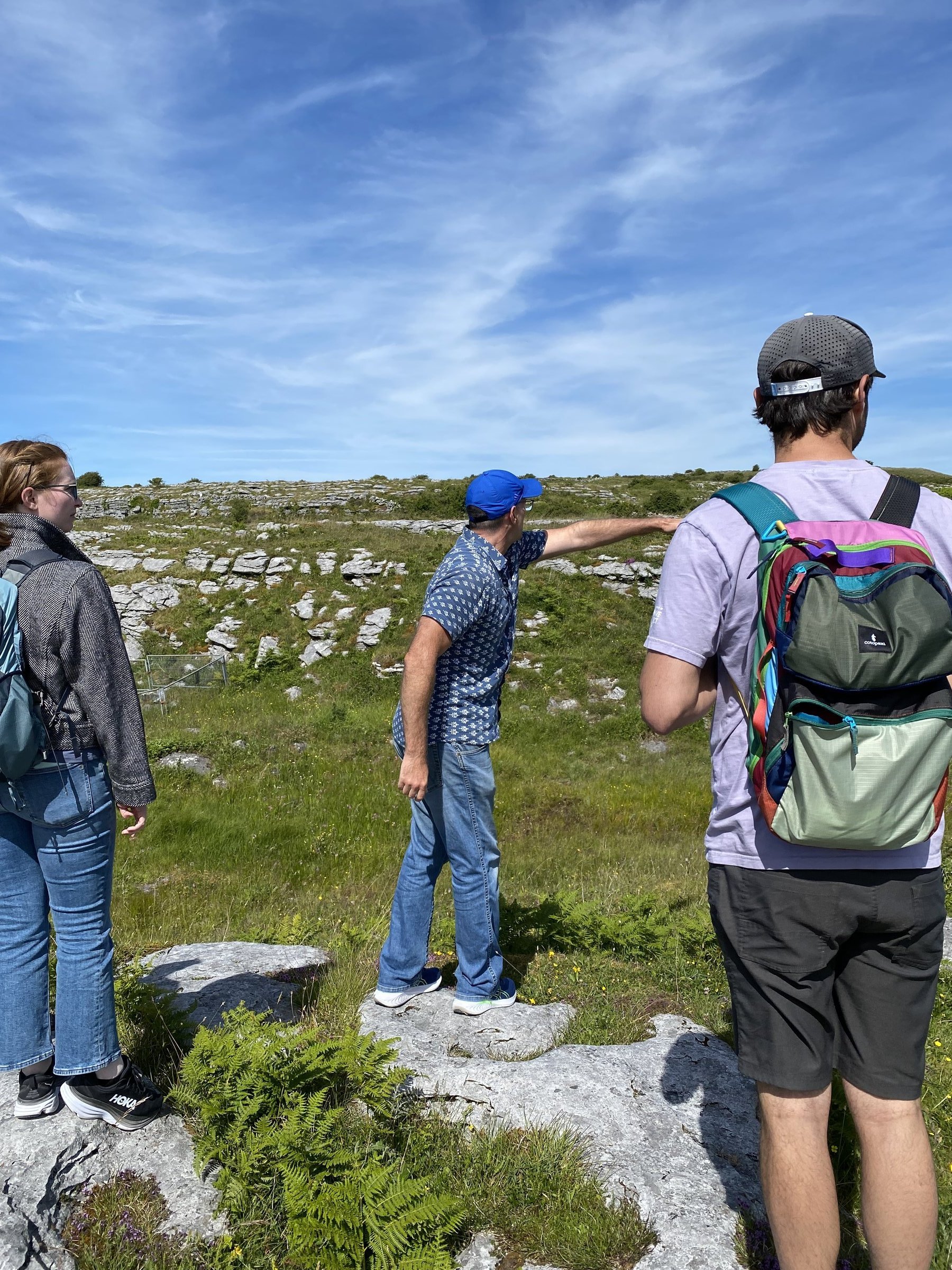 Three hikers pause on limestone pavement as one man in a blue cap points toward the open landscape ahead.
