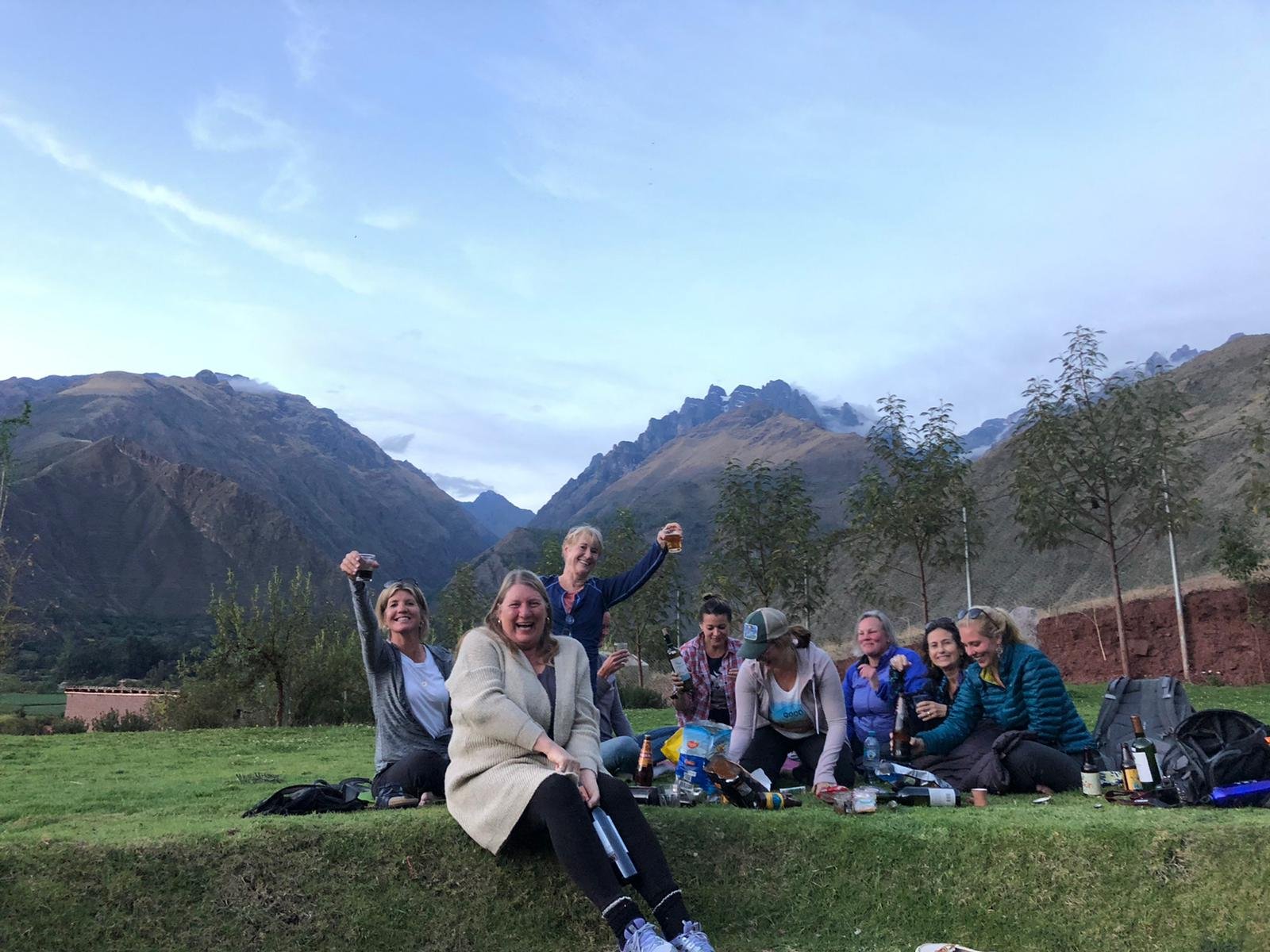 A group of women toast and enjoy a picnic on a grassy hillside with jagged Andes peaks rising behind them in the Sacred Valley.