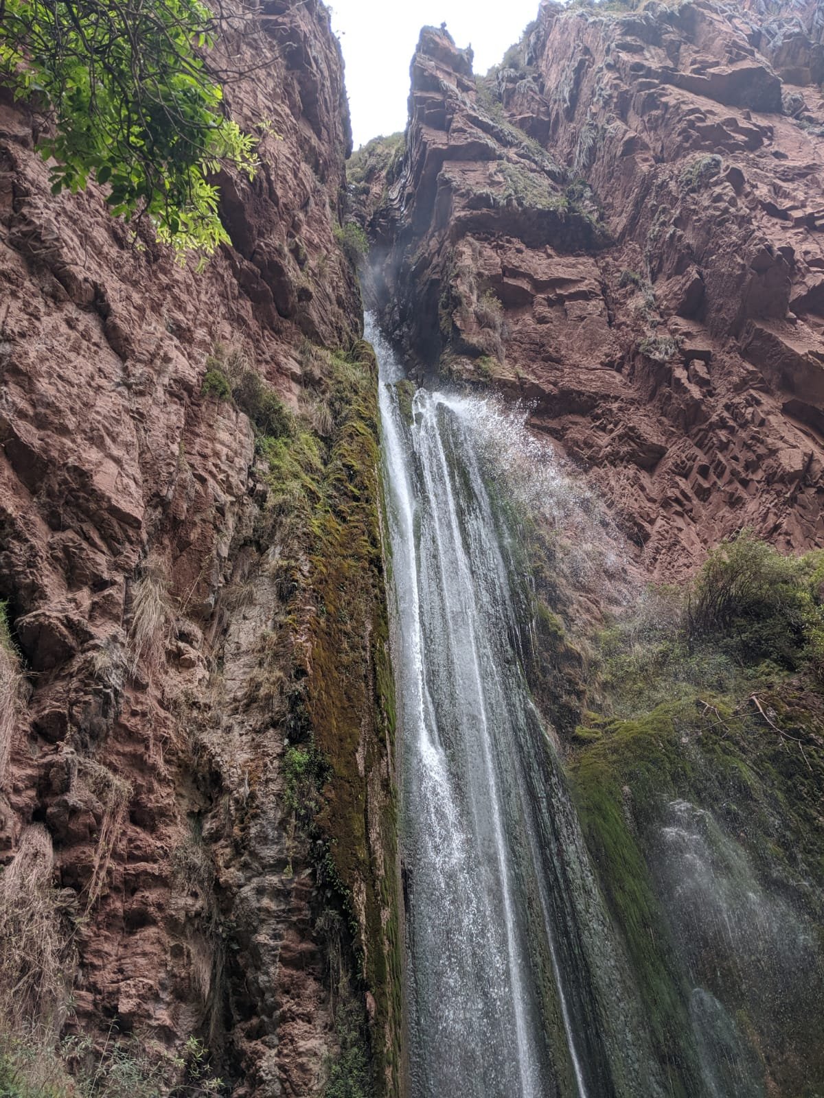 A tall, slender waterfall cascades down a sheer reddish-brown rock cliff in the Sacred Valley.