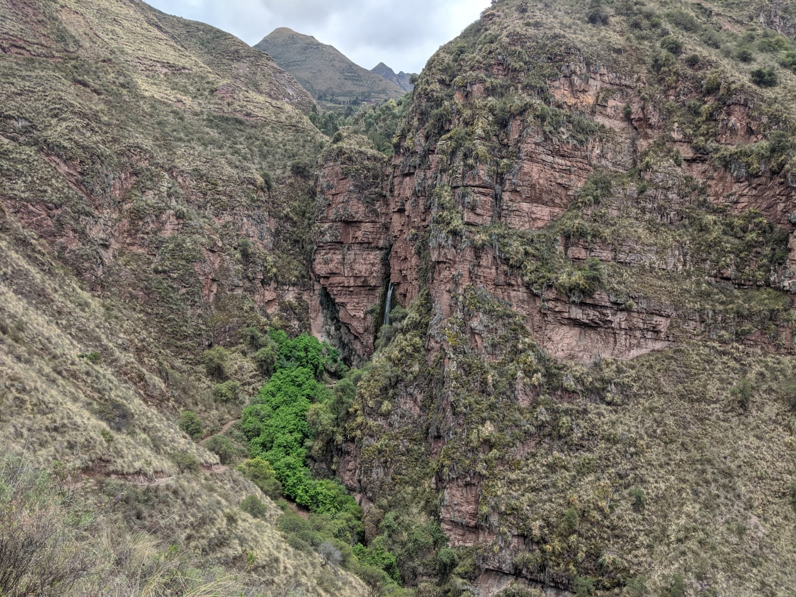 A narrow waterfall drops down a steep rocky cliff into a lush green grove within a mountain canyon in the Sacred Valley.