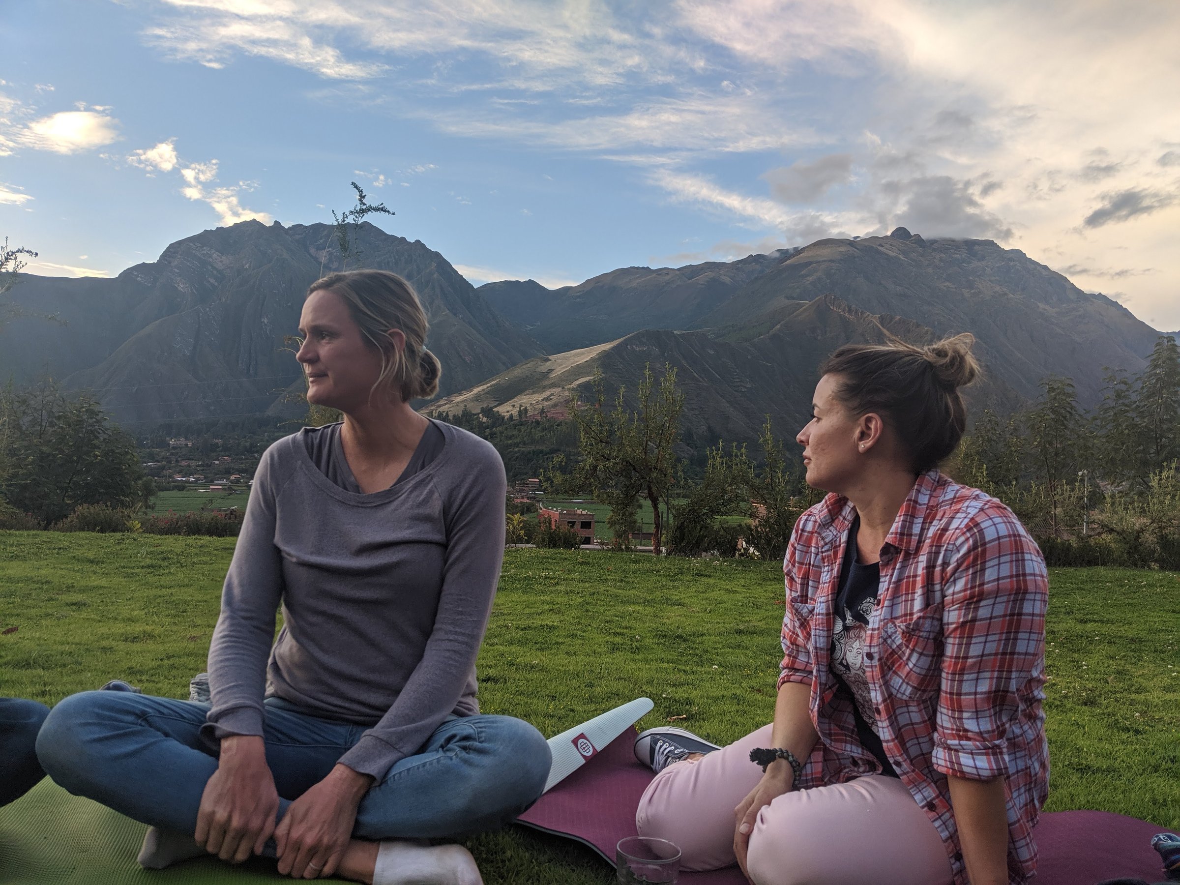 Two women sit on yoga mats in a grassy field with jagged Andes peaks rising behind them in the Sacred Valley.