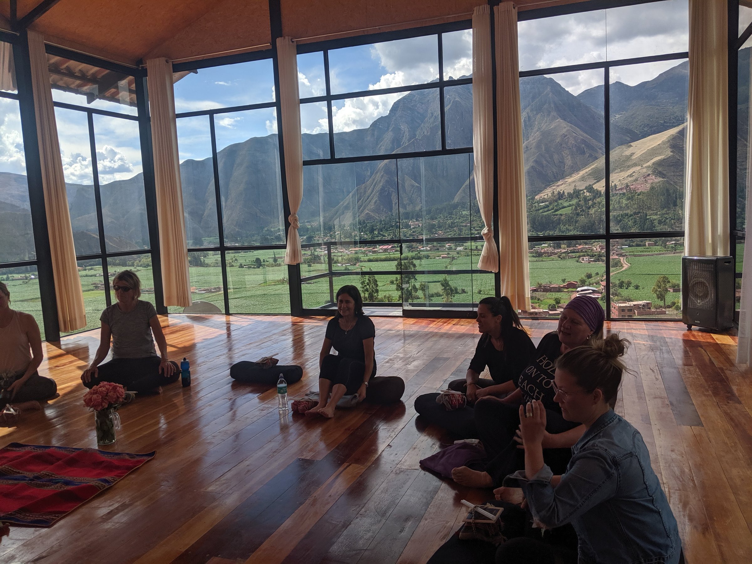 A group of women sits on a polished wooden floor in a bright studio with floor-to-ceiling windows overlooking the lush Sacred Valley and the Andes.
